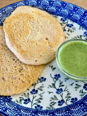 Bunzlau bord met daarop twee Indiase pannenkoekjes van gefermenteerde rode linzen en rijst. Erbij een klein schaaltje met koriander-munt chutney, aangelengd met een beetje sojayoghurt.