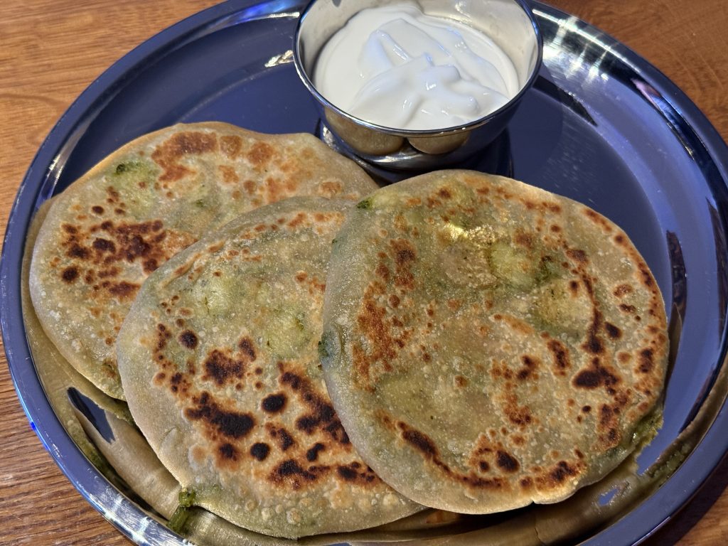 Aan roestvrij stalen bord met daarop een bakje met raita en drie aloo paratha’s