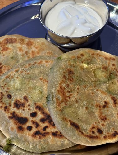 Aan roestvrij stalen bord met daarop een bakje met raita en drie aloo paratha’s